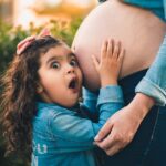A young girl joyfully embraces her pregnant mother's belly in a warm outdoor setting.