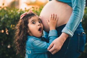 A young girl joyfully embraces her pregnant mother's belly in a warm outdoor setting.