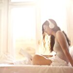 Young happy female with earphones using laptop while sitting on bed in soft daylight by window at cozy bedroom