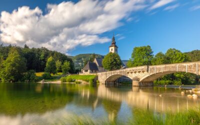 Tranquil view of a stone bridge and historical church over a serene lake in Bohinjska Češnjica, Slovenia.