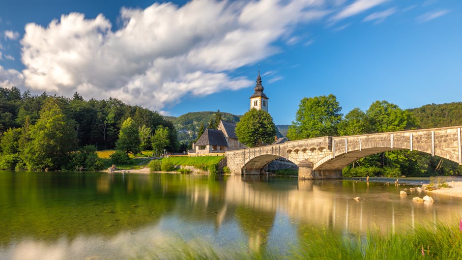 Tranquil view of a stone bridge and historical church over a serene lake in Bohinjska Češnjica, Slovenia.