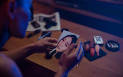 A person holds a photo in a dimly lit room, surrounded by objects suggesting mystery.