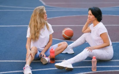 Full length of confident young ladies in sportswear speaking while having break on court after exercising with water bottles near basketball in daylight