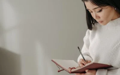 Asian woman writing in a notebook while wearing a knitted sweater indoors, serious and concentrated.