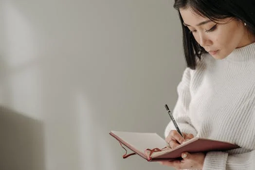 Asian woman writing in a notebook while wearing a knitted sweater indoors, serious and concentrated.