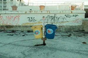 Graffiti-covered wall with blue and yellow recycling bins on a sandy beach.