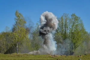 A powerful explosion creates a huge cloud of smoke against a backdrop of green trees, illustrating nature vs. destruction.