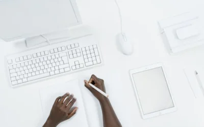 Top-down view of hands writing at a clean, minimalist workspace with a computer and tablet.