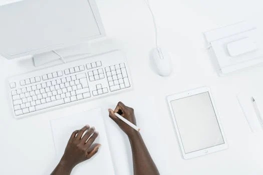 Top-down view of hands writing at a clean, minimalist workspace with a computer and tablet.
