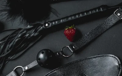 A set of black leather BDSM accessories featuring a ball gag and whip, contrasted with a fresh strawberry.