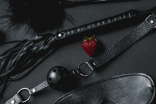 A set of black leather BDSM accessories featuring a ball gag and whip, contrasted with a fresh strawberry.