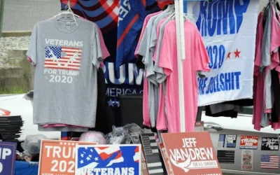 A stall displaying Trump 2020 merchandise including shirts and signs at an outdoor market.