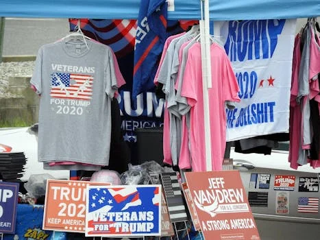 A stall displaying Trump 2020 merchandise including shirts and signs at an outdoor market.