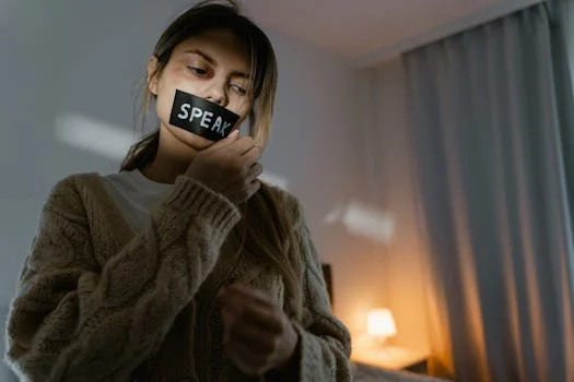 A woman standing in a dimly lit room removing tape with 'SPEAK' written, symbolizing empowerment.