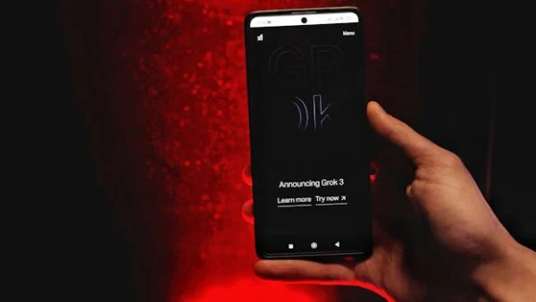 A hand holds a smartphone displaying Grok 3 announcement against a red background.