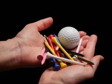 Hands holding a collection of colorful golf tees and a golf ball against a black background.