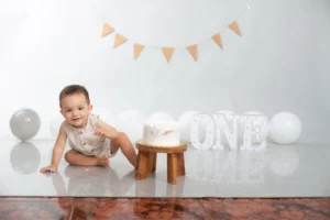 Smiling baby with birthday cake and decorations in a festive indoor setting.