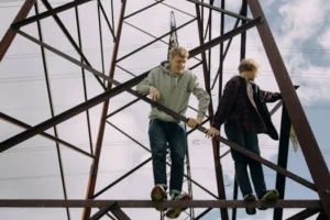 Two teenagers climb a power line tower, enjoying a daring outdoor adventure against a sunny sky.