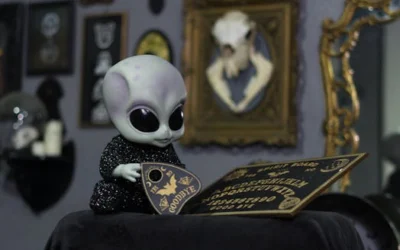 Alien doll interacting with an Ouija board in a decorated room with vintage frames.