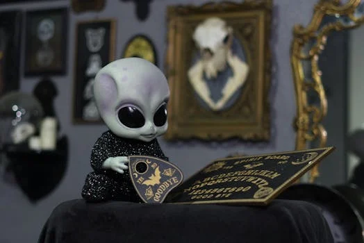 Alien doll interacting with an Ouija board in a decorated room with vintage frames.