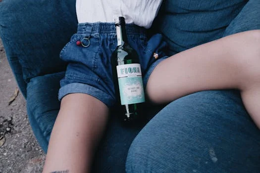 Young person relaxing on blue chair with denim shorts and green wine bottle.