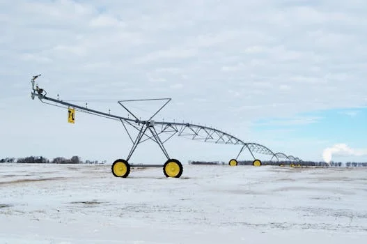 A snow-covered agricultural field with a large metal irrigation sprinkler system under a cloudy sky.