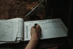 Close-up of a person writing in an outdoor logbook on a table.
