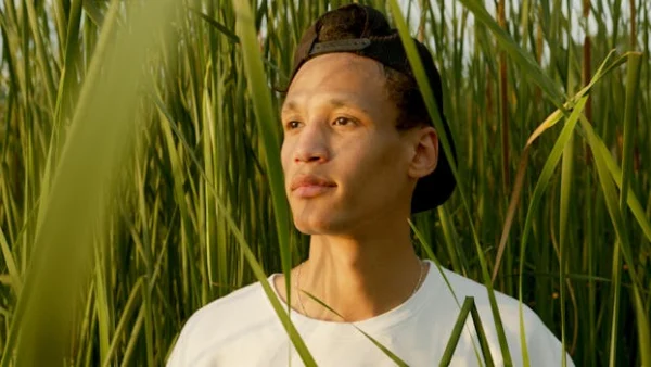 A young man stands peacefully among tall grass in a sunlit meadow, embodying relaxation.