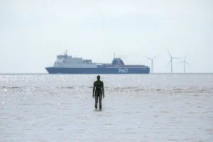 Statue on Crosby Beach, England, facing ship and wind turbines on a bright day.