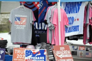 A stall displaying Trump 2020 merchandise including shirts and signs at an outdoor market.